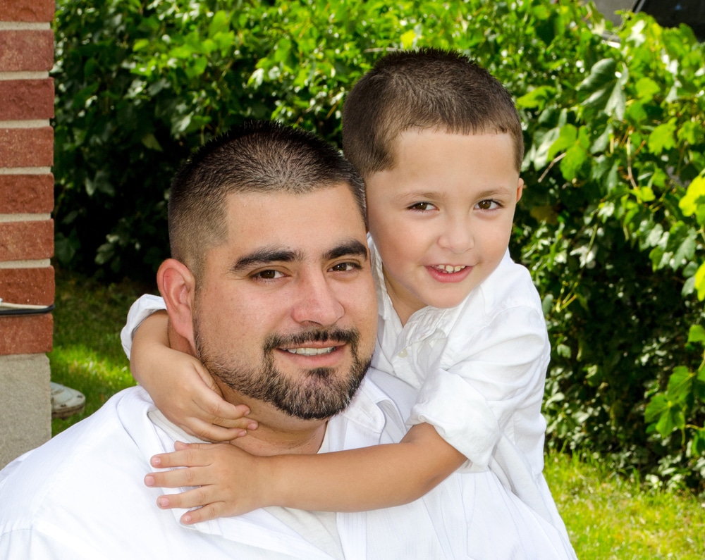 A son hugs his father around the neck, both wearing white shirts outside.